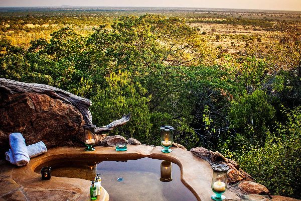 The foreground has a concrete bath filled with water set into a rock on a hillside with a view into the distance across African savannah