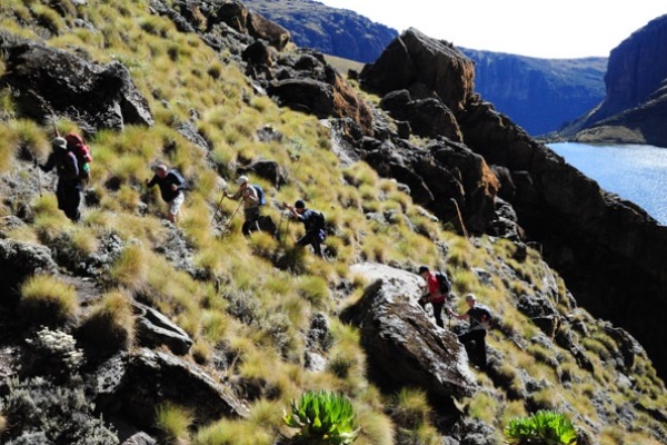 Six climbers with backpacks and walking poles climb a relatively steep path through rocks and grassy tussocks. In the background is a lake.