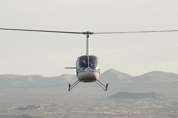 A small helicopter flies directly towards the camera which appears to be at the same height as the helicopter. The pilot is visible in the front window. Below and in the distance is African plain and hills