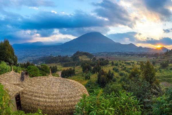 The thatched roofs of Bisate Lodge in the foreground. Bushes and farmed areas in the middle distance. a cone shaped volcano in the distance with the sun just above the horizon and a cloudy sky