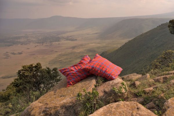 Two cushions covered in traditional Masai red plaid fabric sit in the foreground on a granite rock. Behind them the wall of the Ngorongoro Crater fall away to the crater floor in the mid distance and the far wall of the crater in the distance 