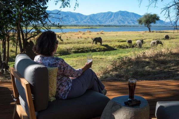 A woman sits looking away from the camera with a drink in her hand on a comfortable chair on a wooden deck. Another drink and a pair of binoculars or on a small table beside her. In the mid distance a herd of elephants grazes on the flood plain of a river. The river and an escarpment are in the background.