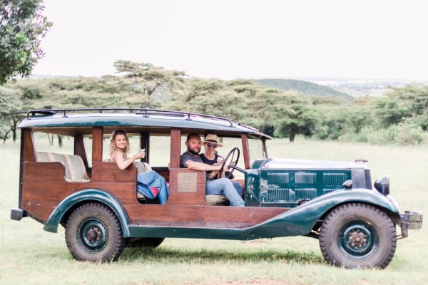 Three people sitting in a vintage car with wooden panelling and three sets of seats. The car is on an open grassy area with trees in the background