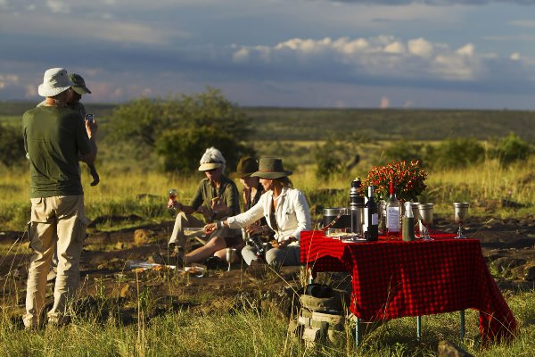 In the foreground is a table with a red tablecloth and drinks. To the left are two men standing. To the centre and slightly behind are three people, perhaps two women and a man, sitting on the ground with drinks in their hands. Behind them are the African plains with grass and shrubs