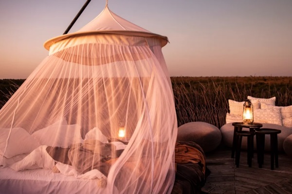 A double bed on an open air platform with a mosquito net suspended above it. It is dusk and there are traditional oil lanterns on tables behind and to the side of the bed