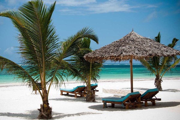 A white sandy beach with the ocean beyond. On the beach are sunbeds, two if which are under a sun umbrella made of palm fronds. There are also three short palm trees on the beach